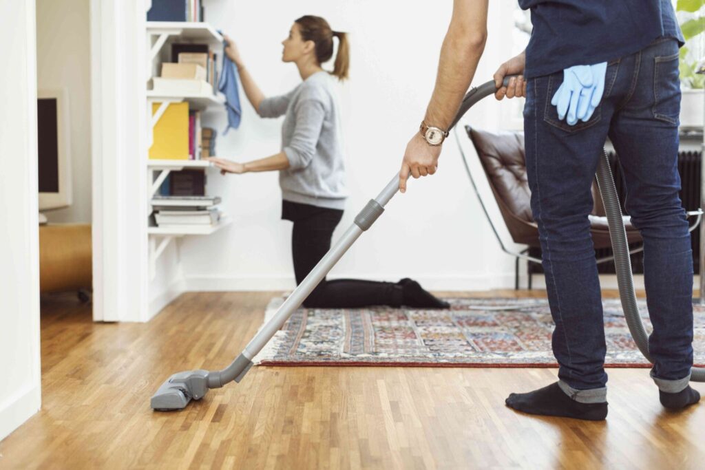 low section of man vacuuming floor while woman cleaning shelves in background at home 562452153 5ac76ae7ae9ab80037f09a24